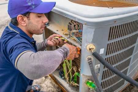 a professional electrician man is fixing the heavy unit of an air conditioner at the roof top of a building and wearing blue uniform and head cap HVAC Home Services SMS